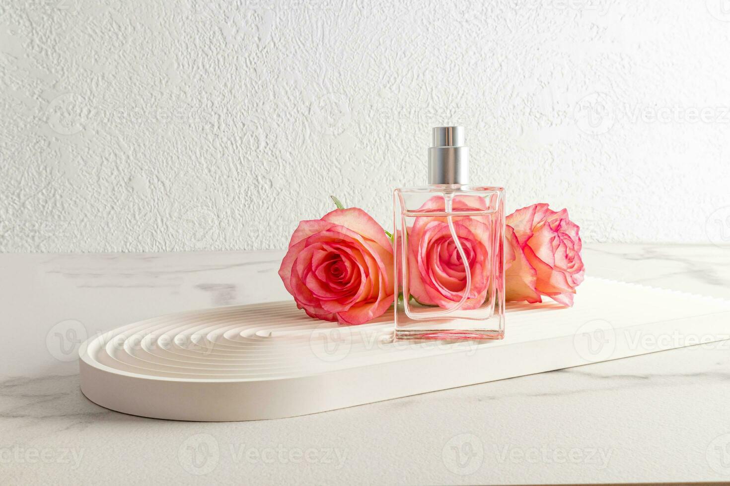 A stylish bottle of perfume against the background of fresh pink roses stands on a white podium in the form of an arch. A modern image of perfume. photo