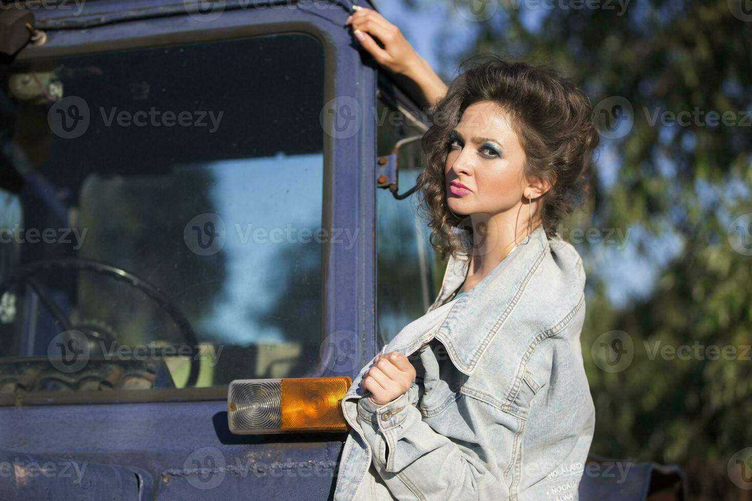 Beautiful young woman in a denim jacket posing against the backdrop of a tractor. Girl in the style of the eighties. photo