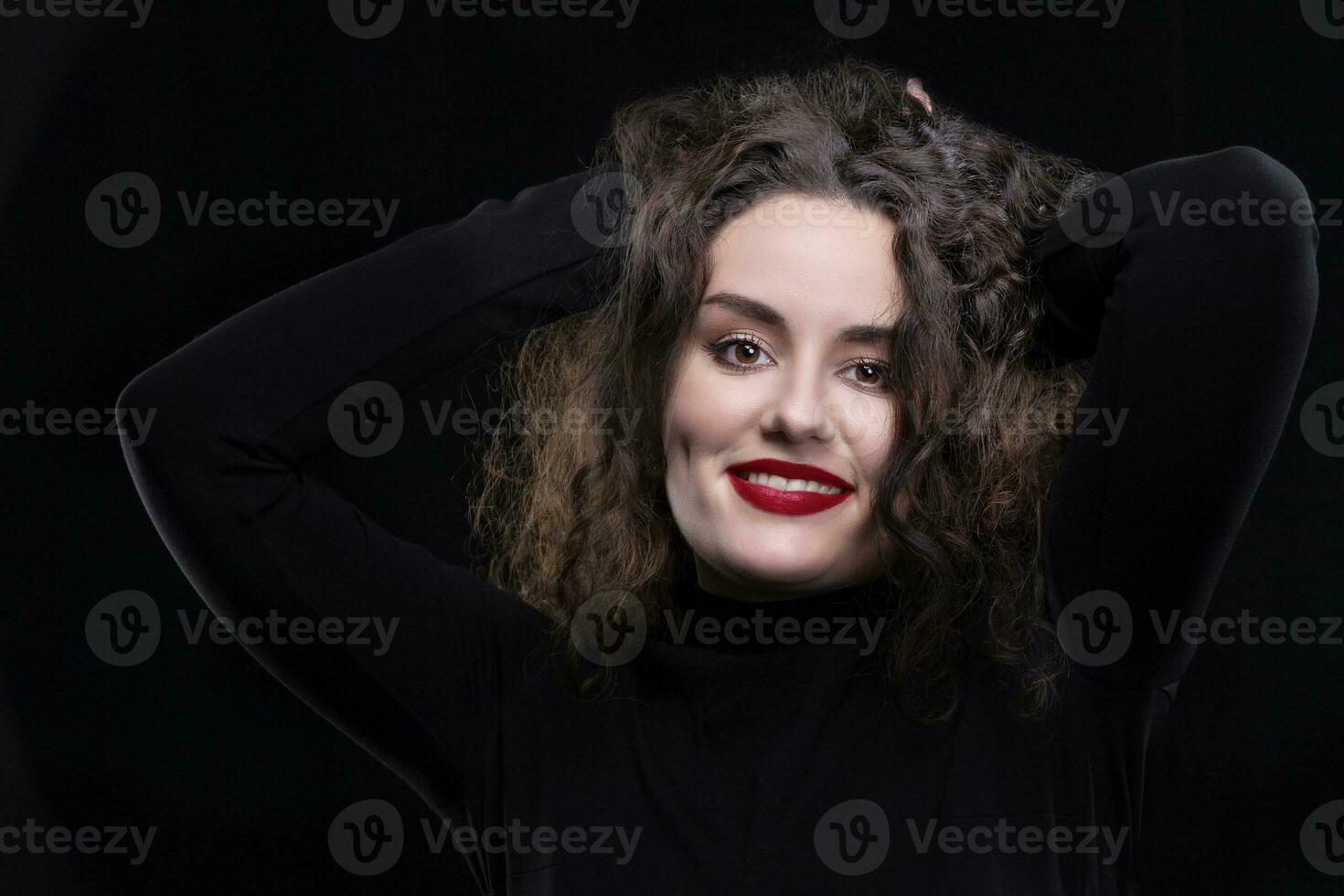 A beautiful brunette girl with curly hair smiles and looks at the camera on a dark background. photo