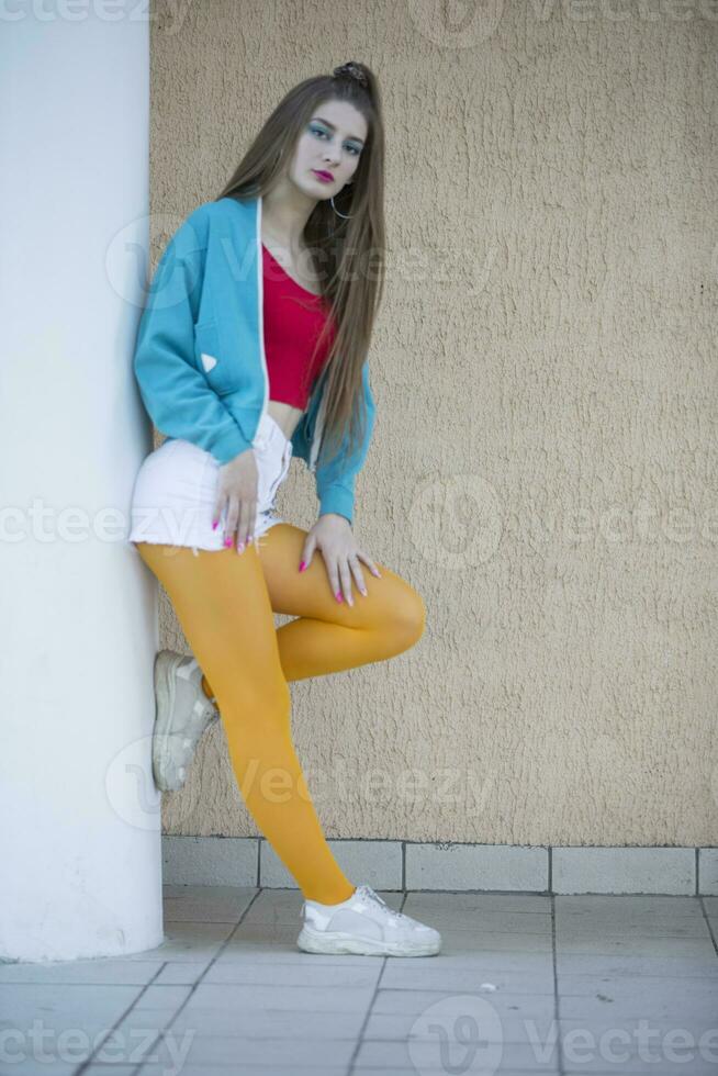 A brunette girl dressed in the style of the nineties of the last century stands against a beige wall. A bright girl in yellow tights. photo