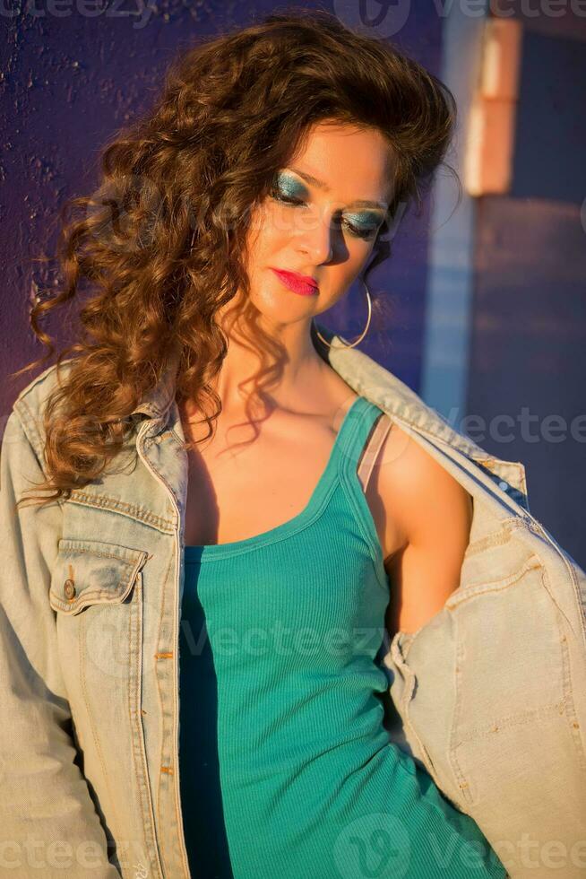 hermosa joven mujer en un mezclilla chaqueta en el antecedentes de un pelado púrpura pared. niña con brillante maquillaje en el estilo de el ochenta en el calle. foto