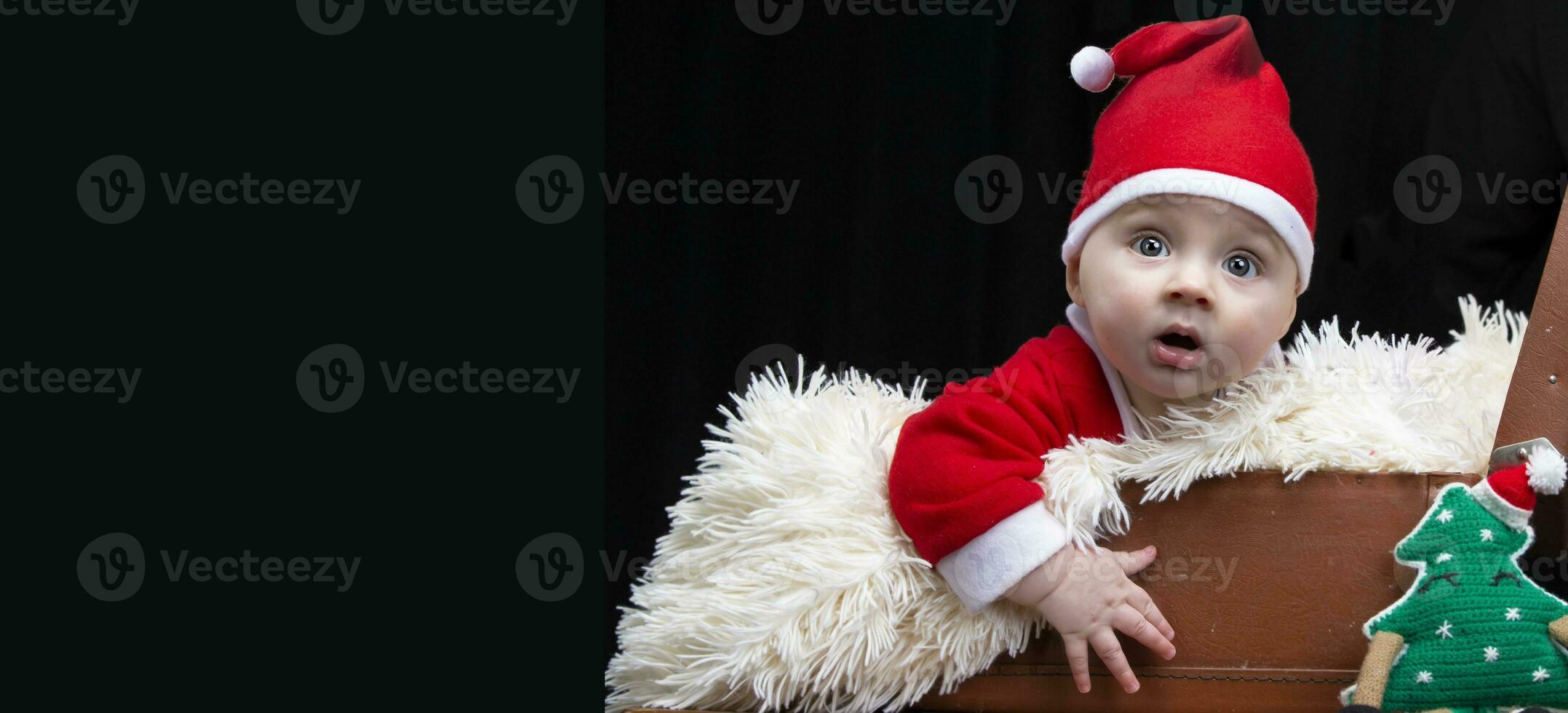 gracioso Navidad niño en un Papa Noel claus sombrero en un retro maleta en un negro antecedentes. niño en Navidad. foto