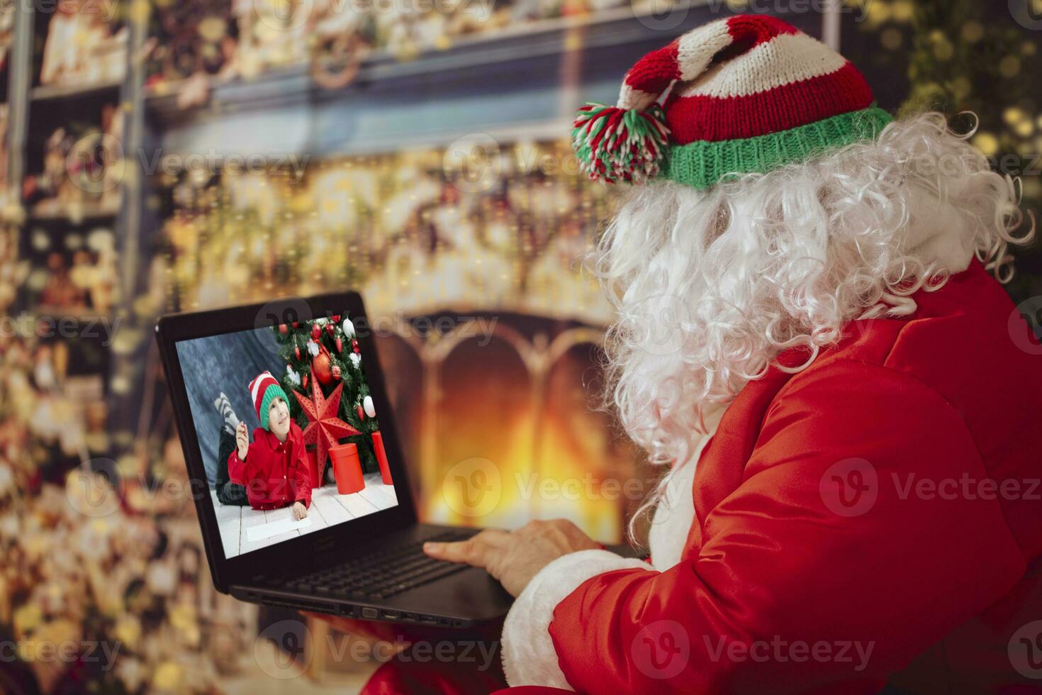 Happy Santa Claus makes video call toddler talking to child, greeting Merry Christmas, Happy New Year in virtual video online chat on laptop, sitting in front of the fireplace. photo