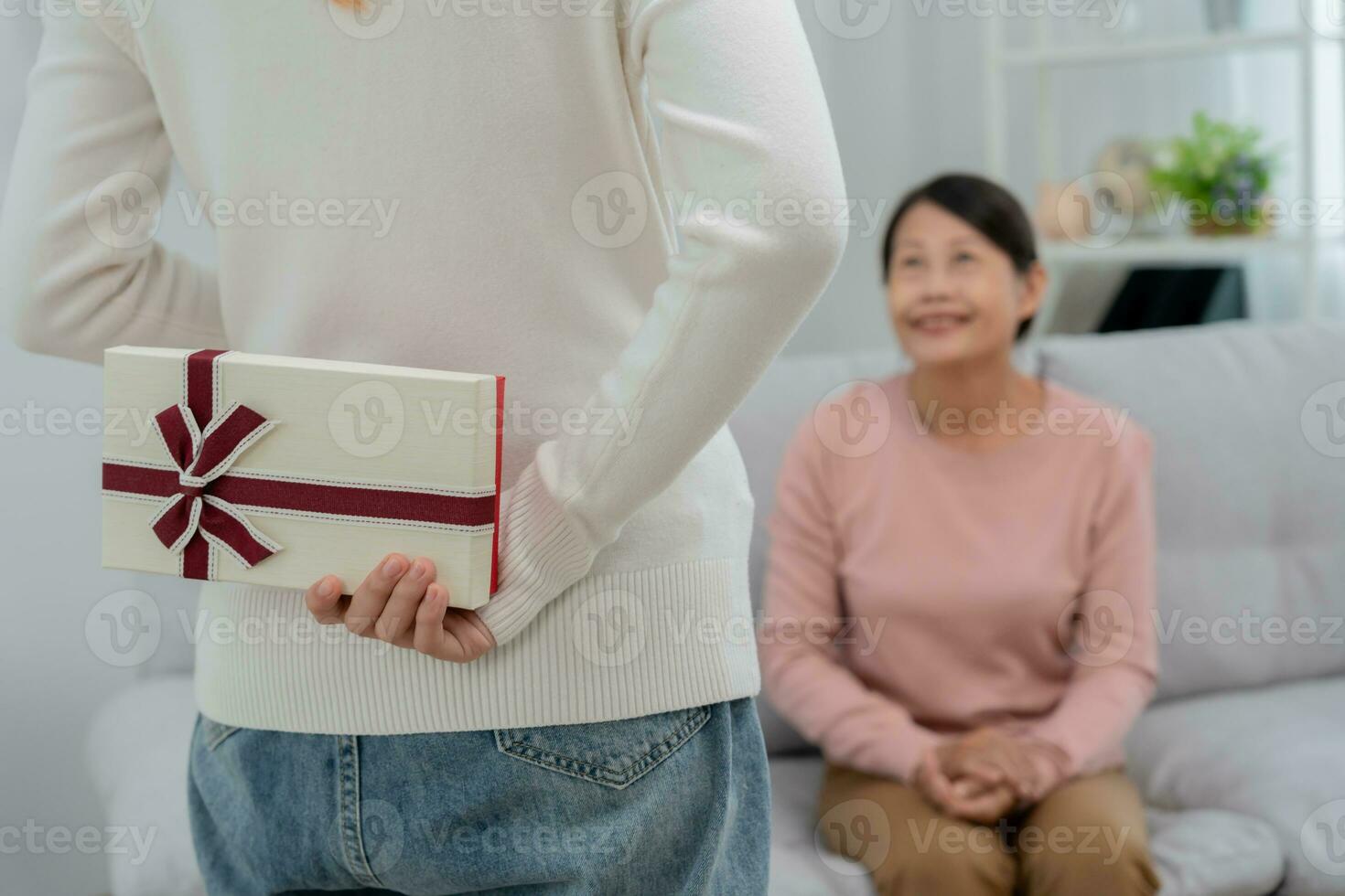 asiático adolescente muchachas son ocultación regalo cajas para su hermosa mamás detrás su espaldas mientras mamá es sentado en el sofá a hogar. celebrando especial ocasión juntos a hogar, contento cumpleaños a mamá foto