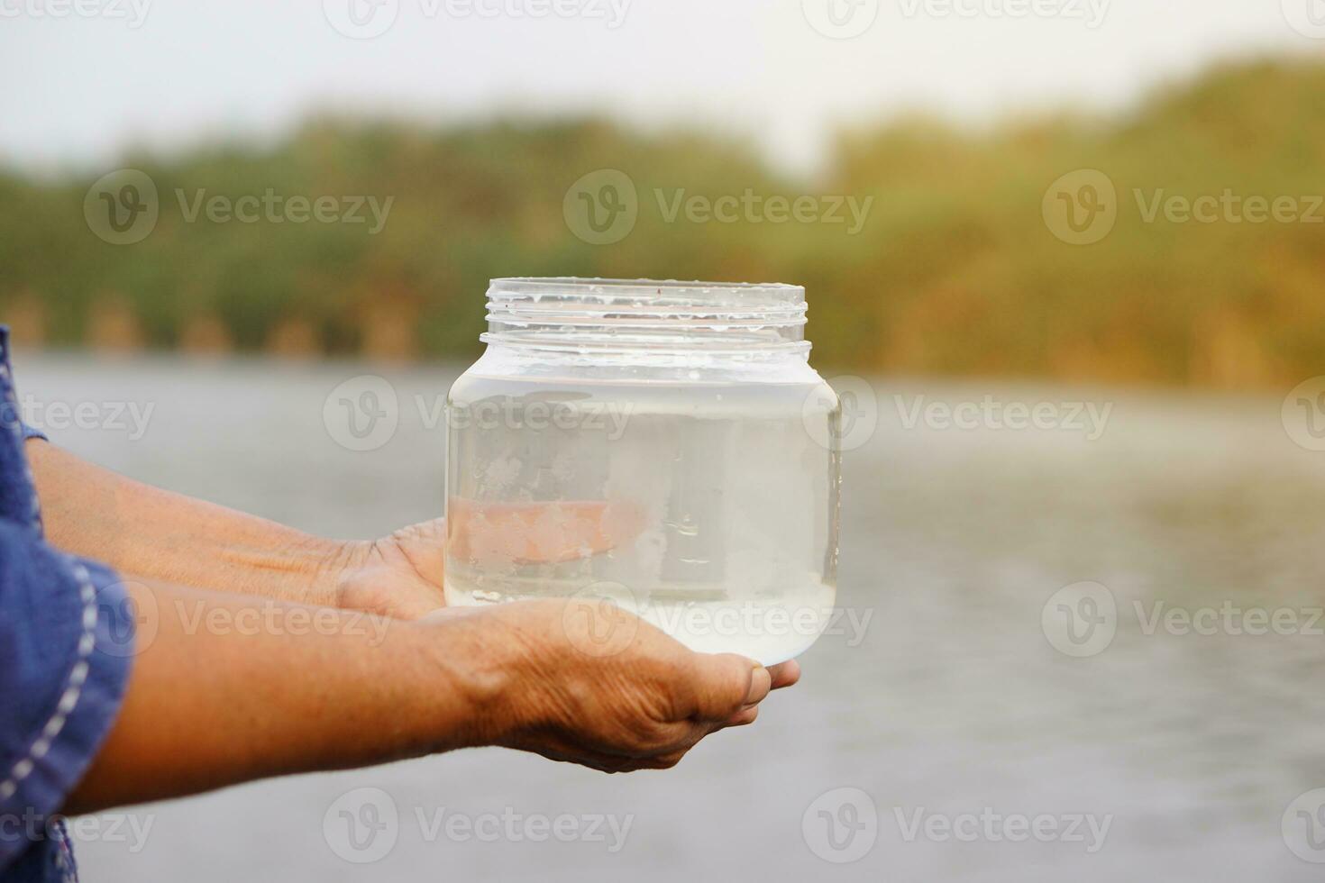 Closeup hands hold transparent jar that contain sample water from the lake. Concept, explore, inspect quality of water from natural source. Ecology survey. Environment conservation. photo