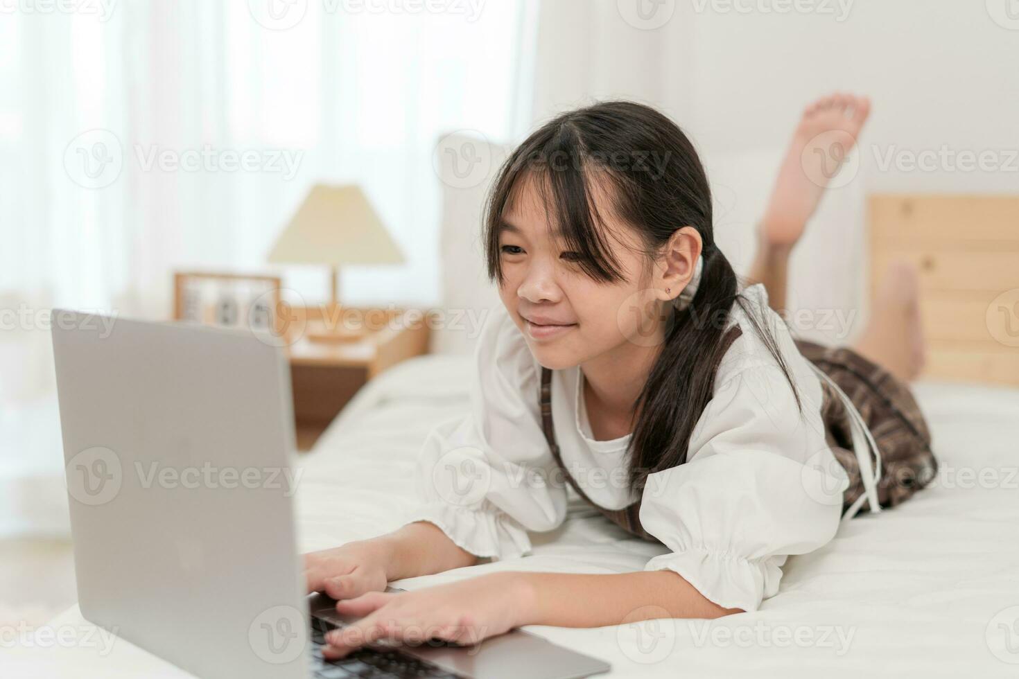 Asian little girl smiling and using a laptop on the bed in the room photo