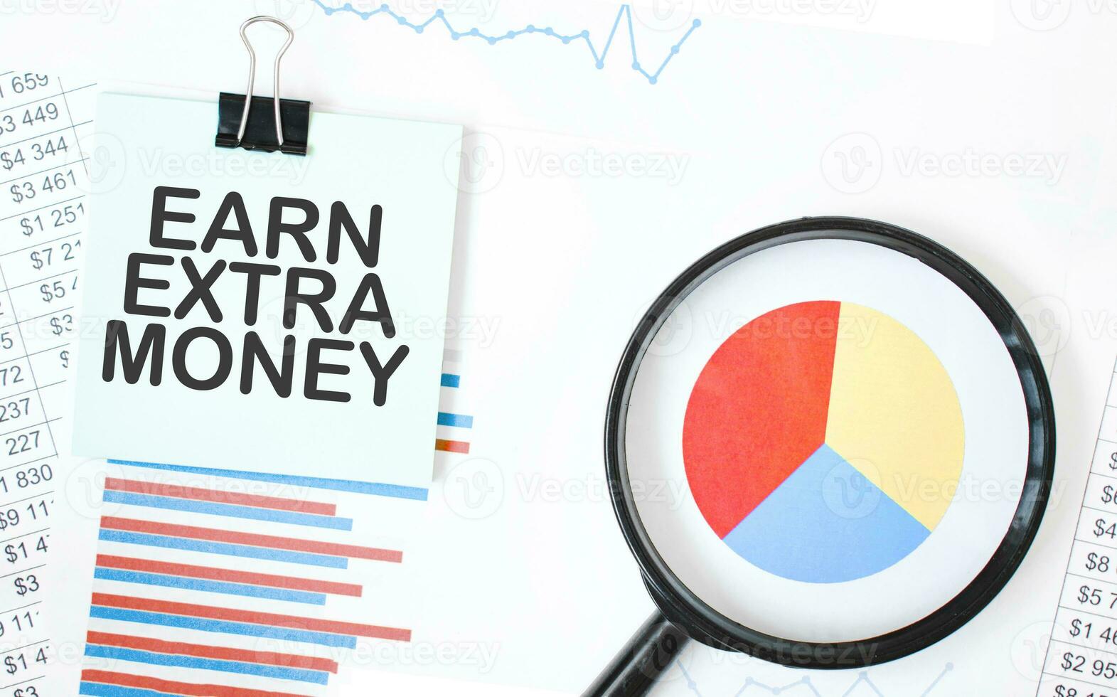 Green card on the white notepad and magnifier on the financial documentation. Text EARN EXTRA MONEY. Business concept photo