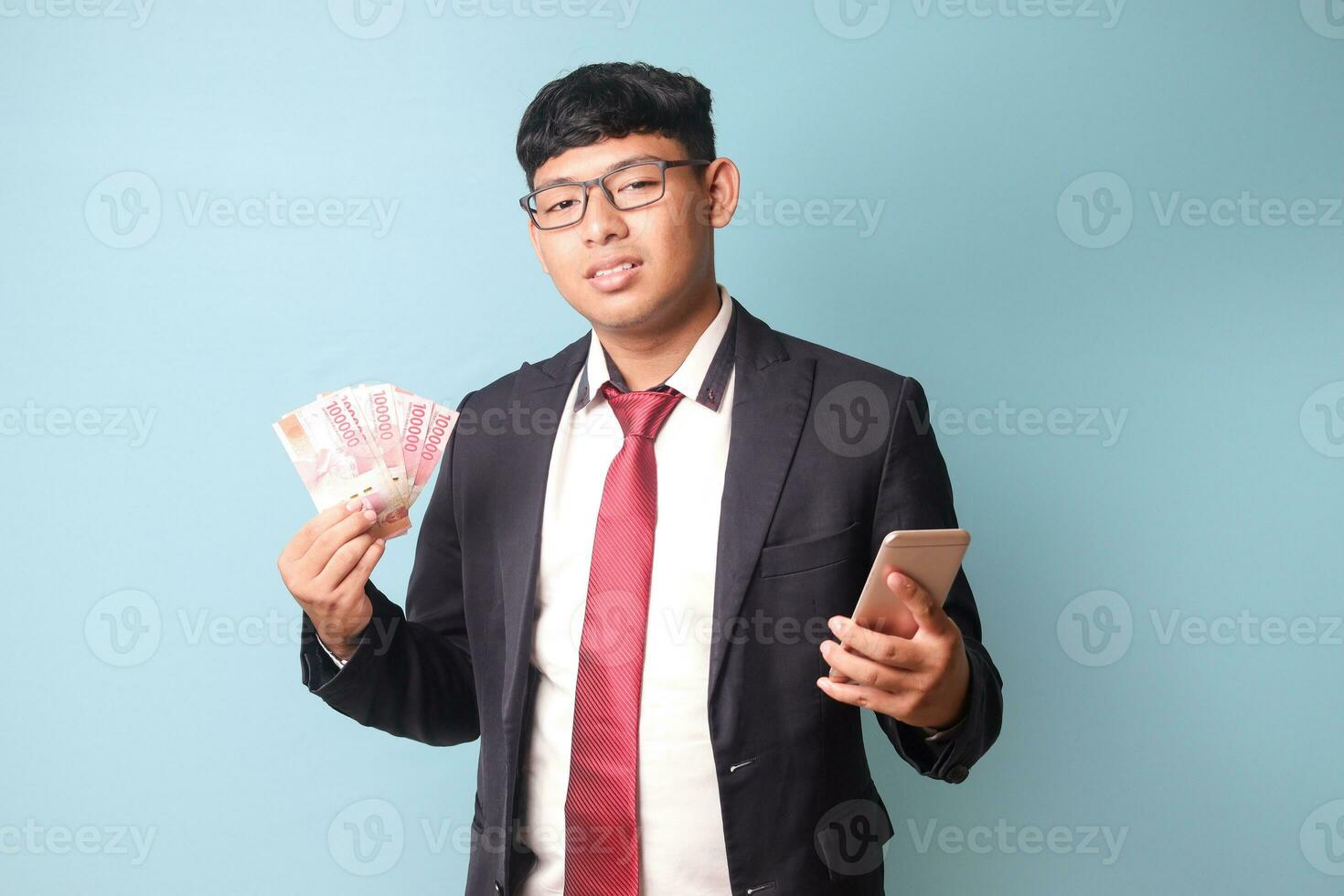 Portrait of young Asian business man in casual suit looking at camera with slight head tilt while holding phone thousand rupiahs. Isolated image on blue background photo