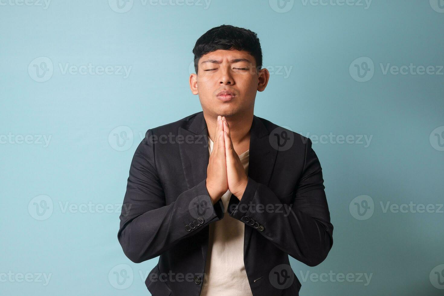 Portrait of young Asian business man in casual suit begging for forgiveness. Isolated image on blue background photo