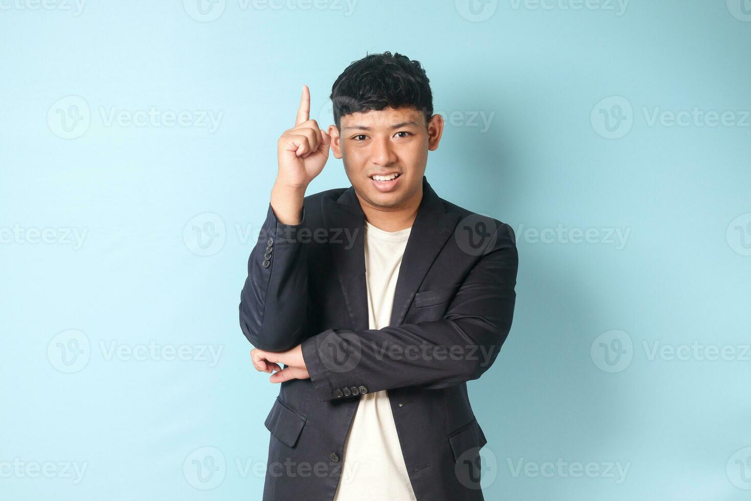 Portrait of young Asian business man in casual suit pointing upward while looking at camera.Isolated image on blue background photo