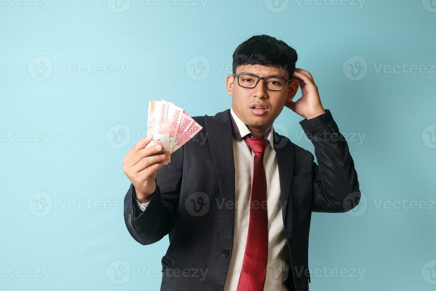 retrato de joven asiático negocio hombre en casual traje escarchando cabeza mirando confuso mientras participación mil rupias. aislado imagen en azul antecedentes foto