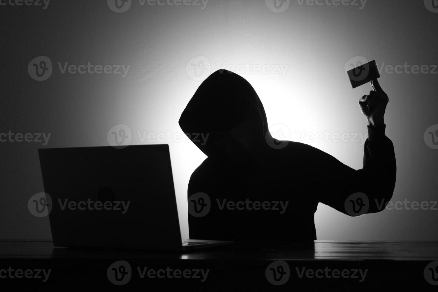 misterioso hombre vistiendo negro capucha utilizando ordenador portátil y participación crédito tarjeta en el oscuro habitación. hacker y ciber seguridad concepto. foto