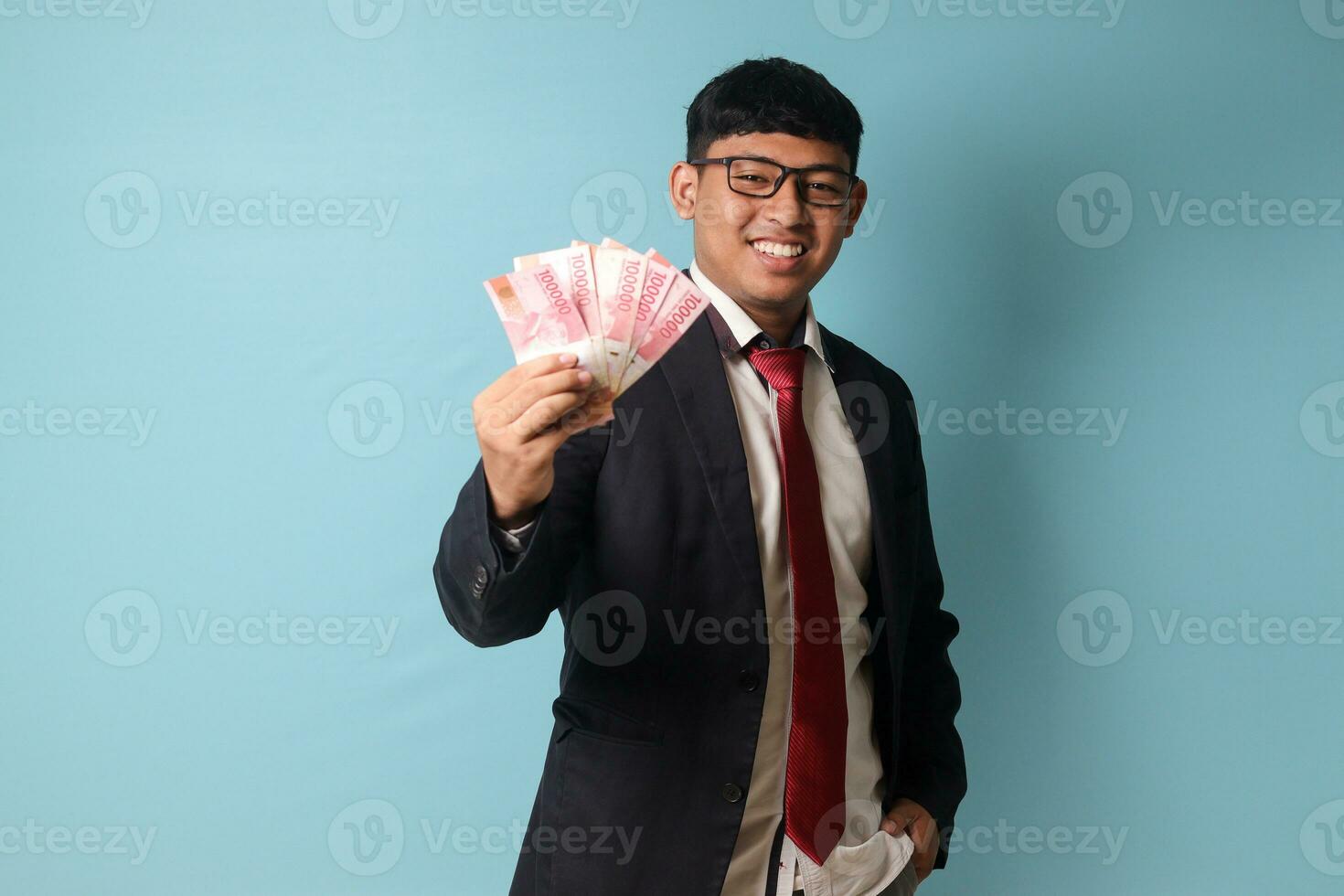 Portrait of young Asian business man in casual suit smiling while holding thousand rupiahs proudly. Isolated image on blue background photo