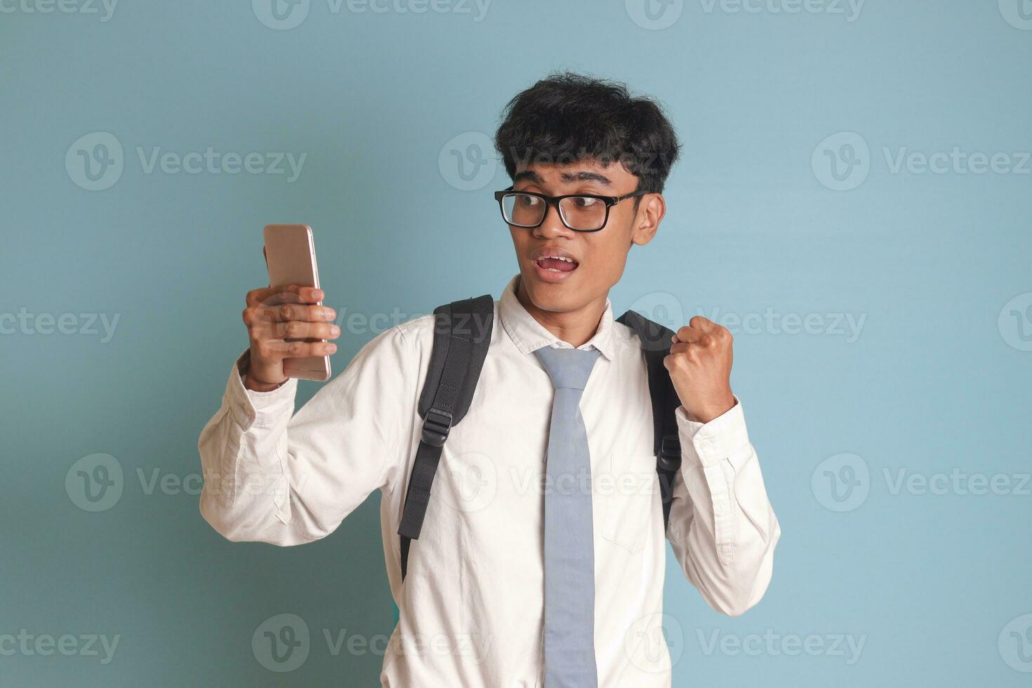 Indonesian senior high school student wearing white shirt uniform with gray tie raising his fist, celebrating success while holding a mobile phone. Isolated image on blue background photo