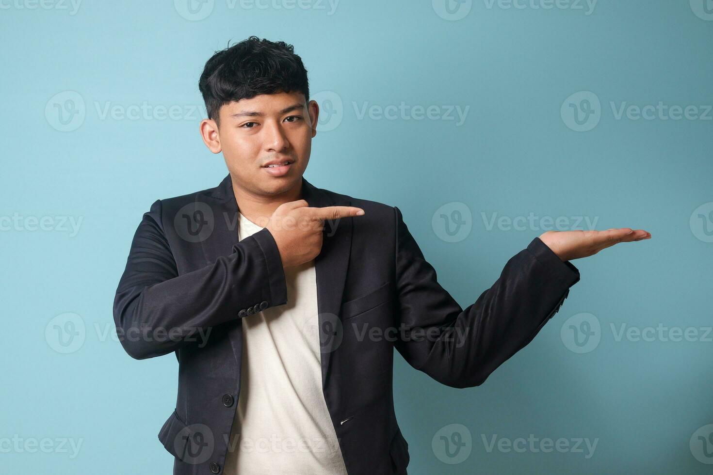 Portrait of young Asian business man in casual suit presenting at something while pointing at it with happy expressions. Isolated image on blue background photo