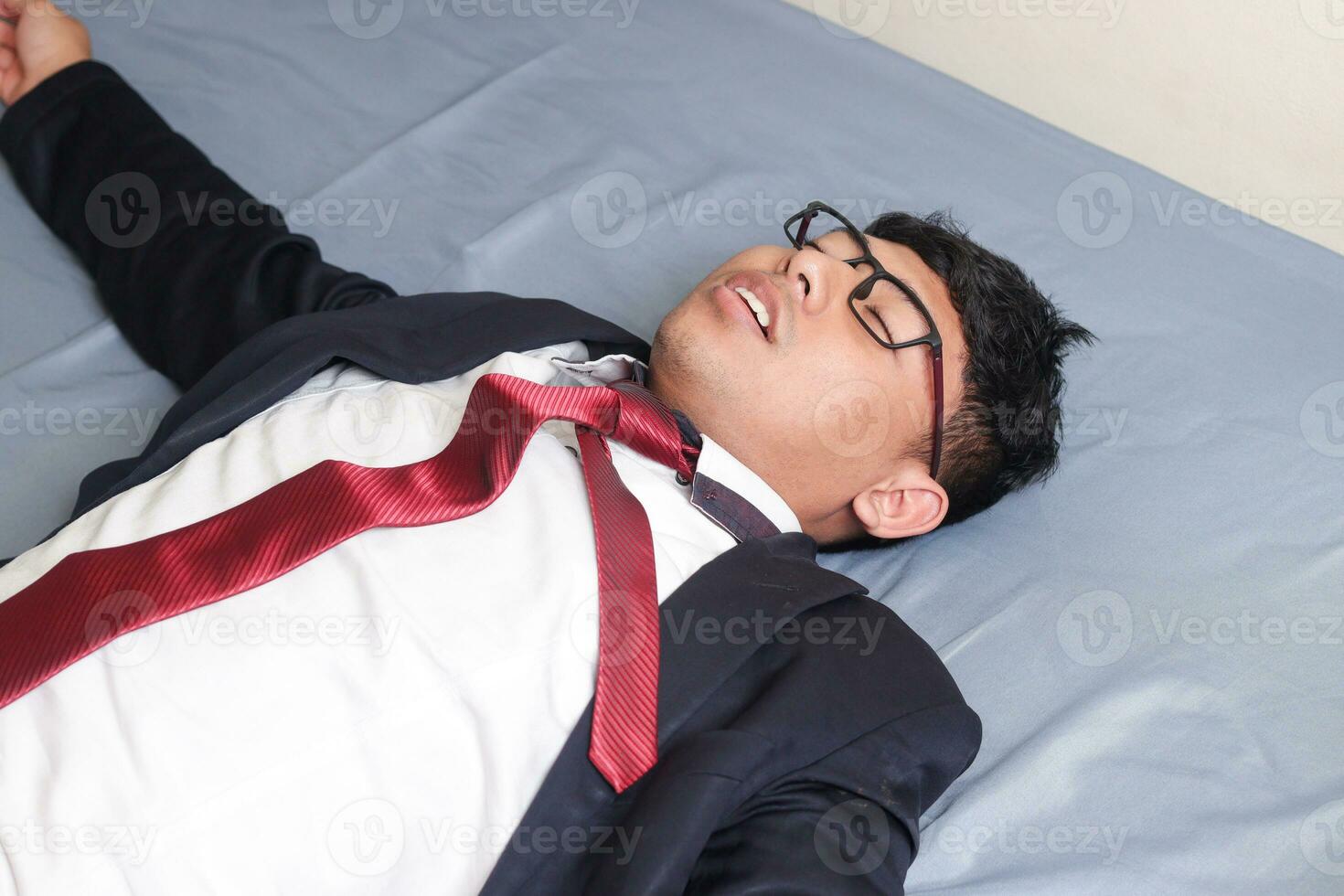 Portrait of young Asian business man in casual suit utterly defeated in bed with very exhausted expressions. Isolated image on blue background photo