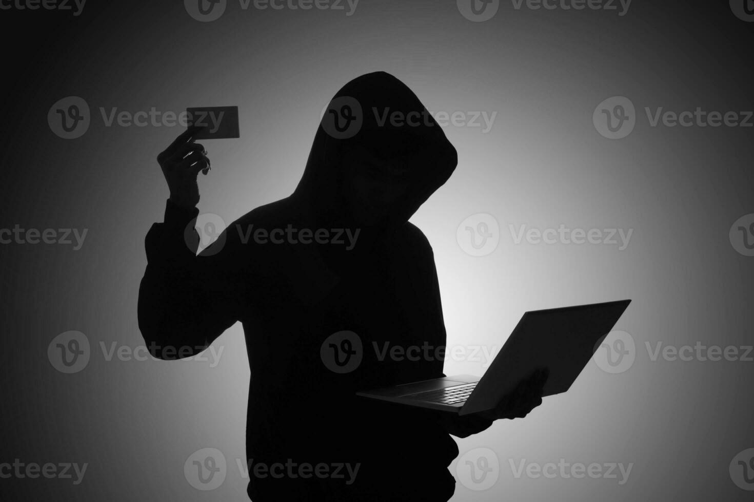 Mysterious man wearing black hoodie using laptop and holding credit card in the dark room. Hacker and cyber security concept. photo