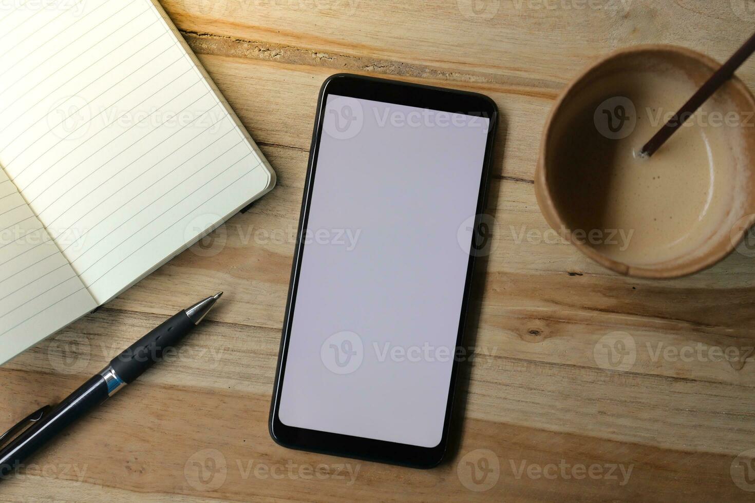 Blank screen mobile phone with empty notebook, pen, and a cup of coffee on wooden table during the day. Office work concept. Flat lay images photo