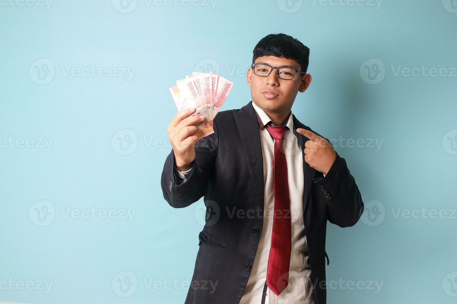 Portrait of young Asian business man in casual suit pointing at thousand rupiahs with serious expressions. Isolated image on blue background photo