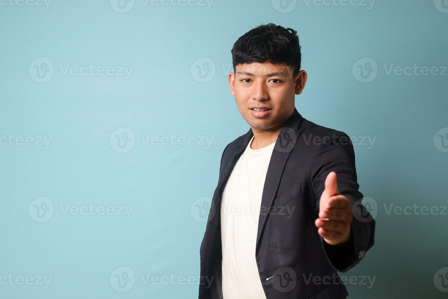 Portrait of young Asian business man in casual suit inviting someone with friendly hand gestures and happy expression. Isolated image on blue background photo