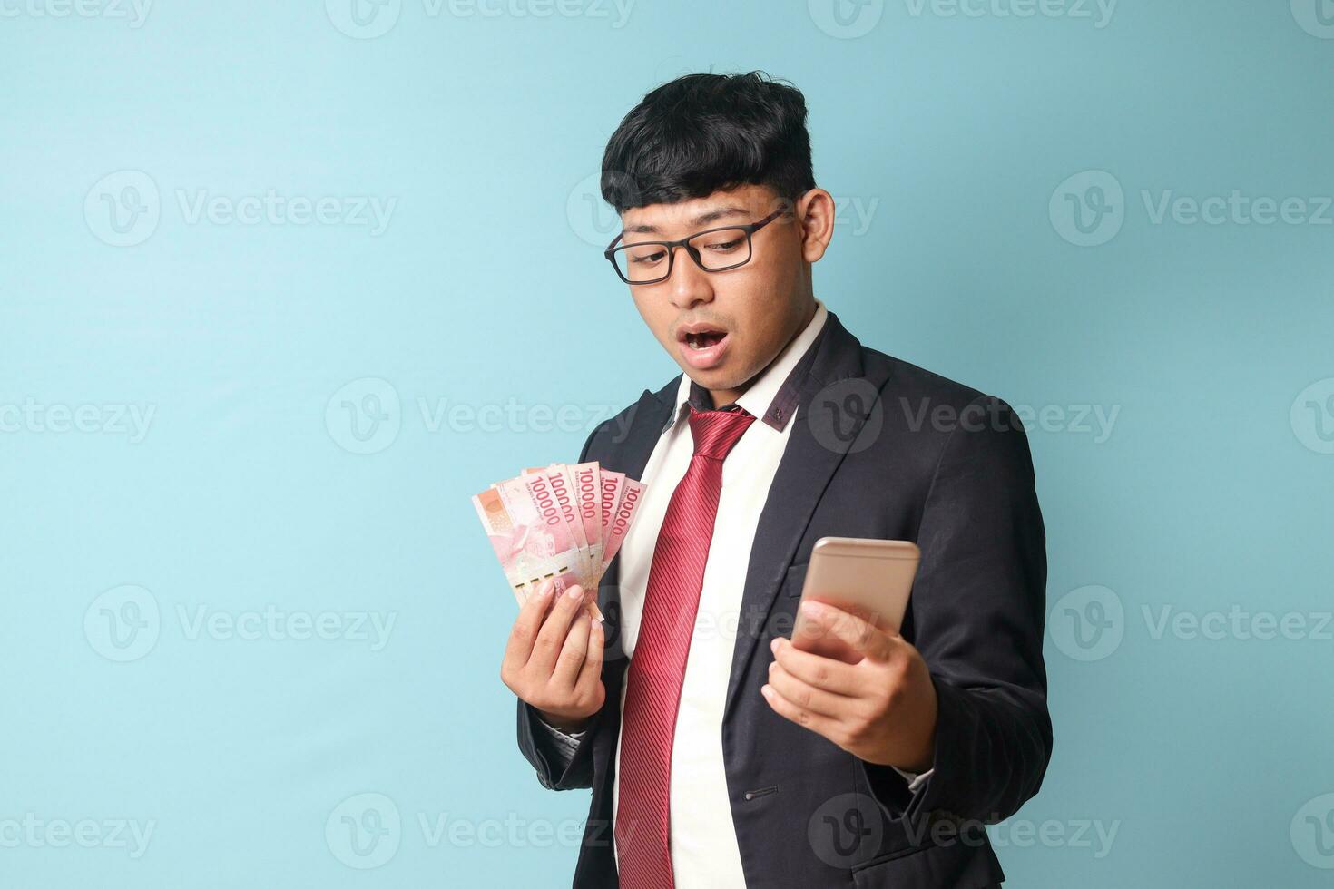 Portrait of young Asian business man in casual suit looking at smartphone surprised while holding thousand rupiahs. Isolated image on blue background photo