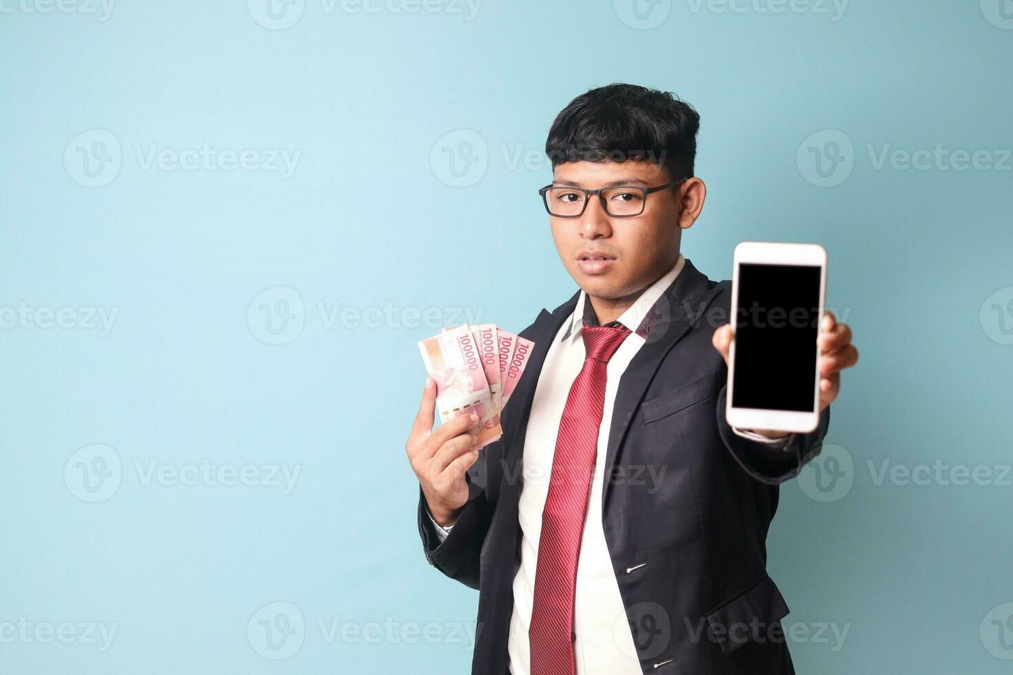 Portrait of young Asian business man in casual suit showing phone and holding thousand rupiahs looking at camera with serious face. Isolated image on blue background photo