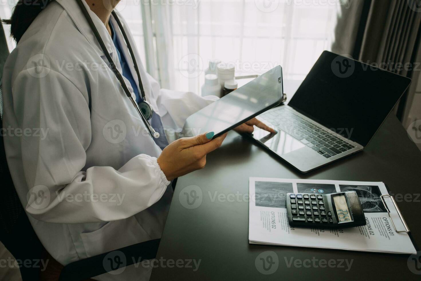 Serious female doctor using laptop and writing notes in medical journal sitting at desk. Young woman professional medic physician wearing white coat and stethoscope working on computer at workplace. photo