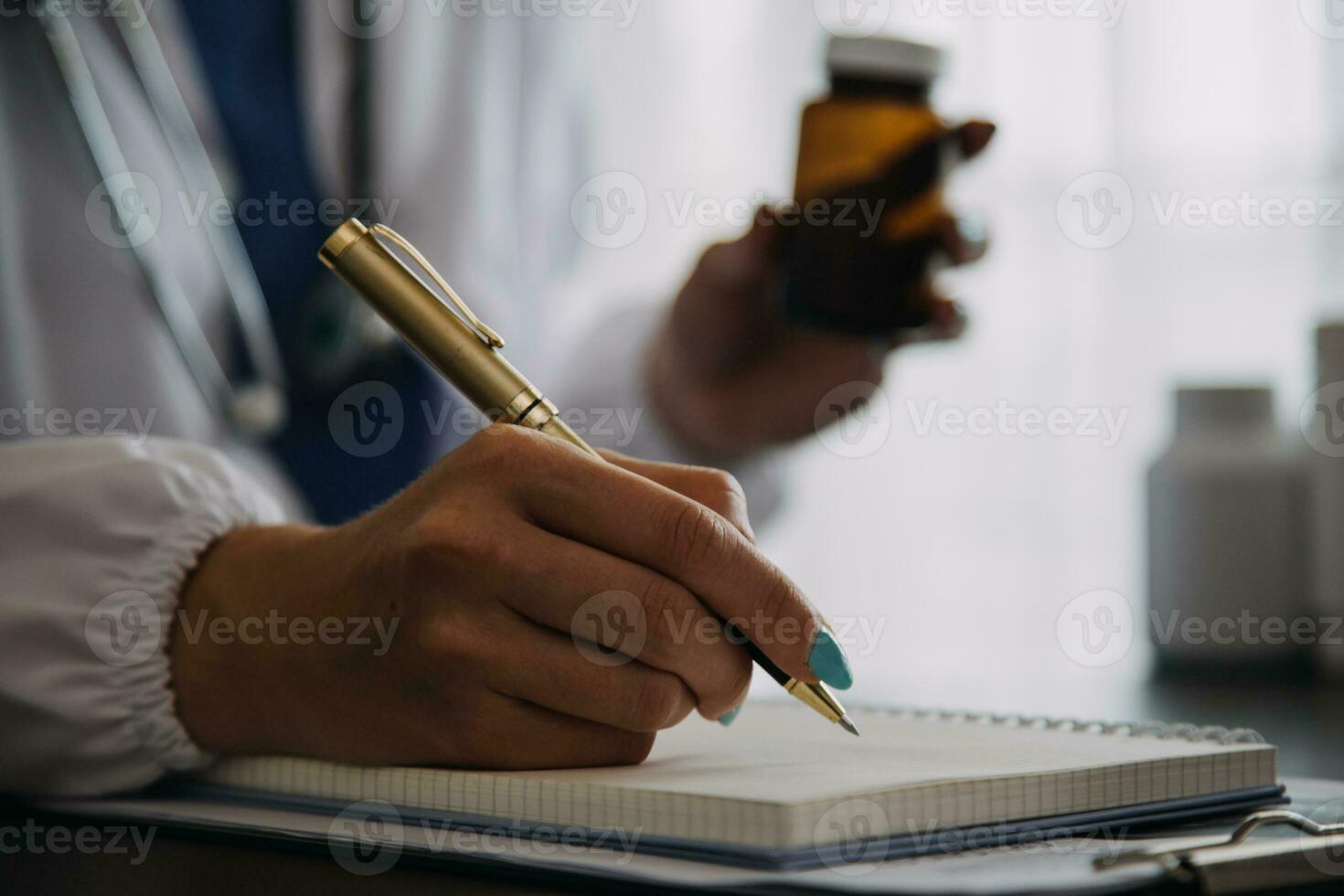 Serious female doctor using laptop and writing notes in medical journal sitting at desk. Young woman professional medic physician wearing white coat and stethoscope working on computer at workplace. photo