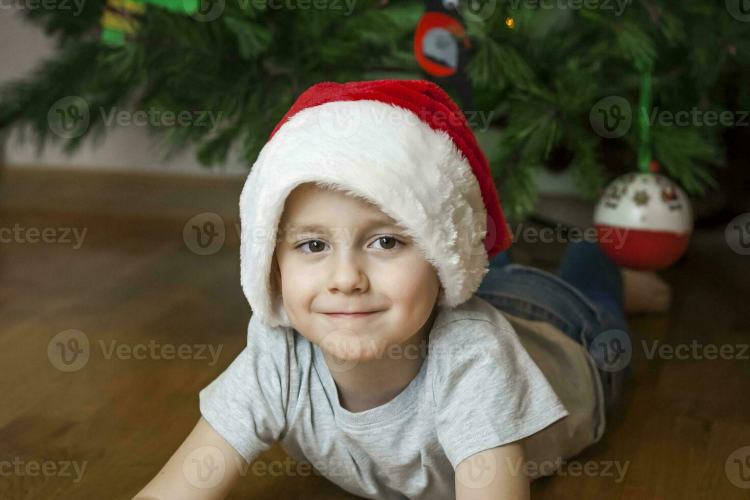 A photo of a beautiful boy in a gray T-shirt and a Santa Claus hat at the Christmas tree, looking into the camera. Portrait in a bright room. Natural, not staged photography.