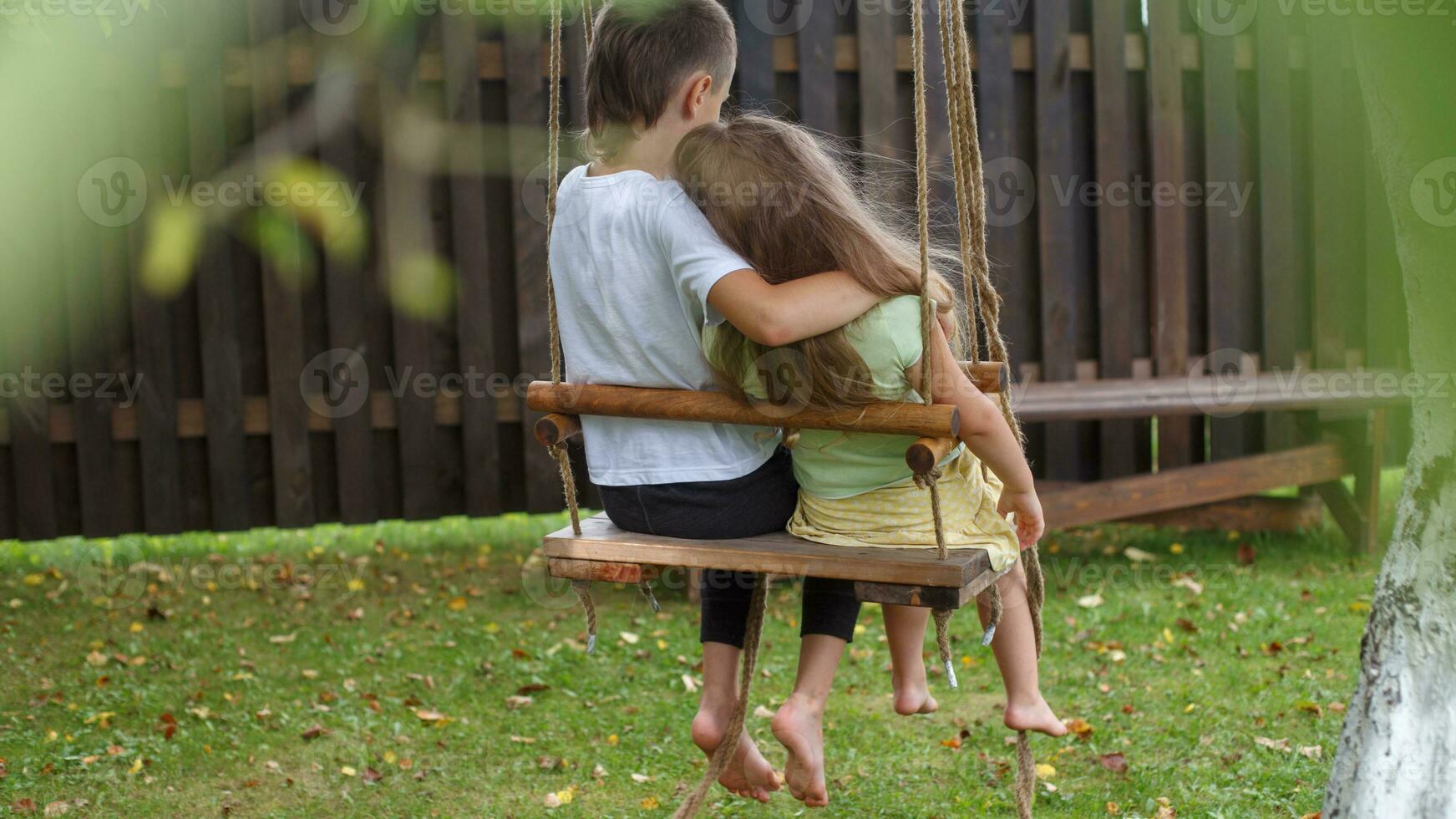 children sitting on a swing in the garden. older brother hugging little sister photo