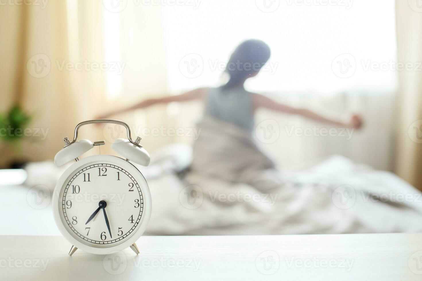 little boy sitting and stretching in bed at home in the morning on a window background with alarm clock photo