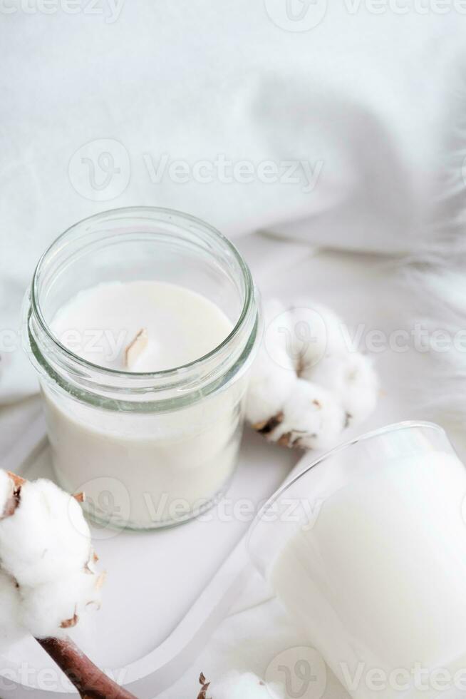 Soy wax aroma candle in white jar on bed with cotton branch. Candle mockup design photo