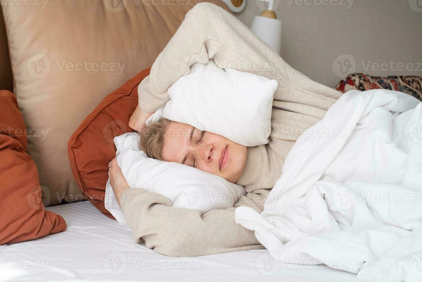 Woman sleeping at bed at home, covering ears with pillows from noise photo