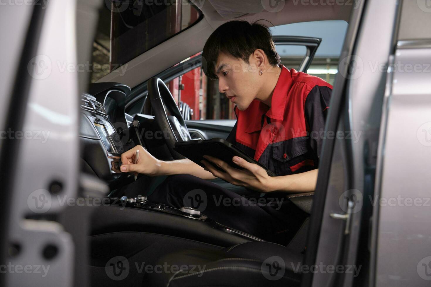 One young expert Asian male automotive mechanic technician checking a maintenance list with tablet in car interior at garage. Vehicle service fix and repair works, industrial occupation business jobs. photo