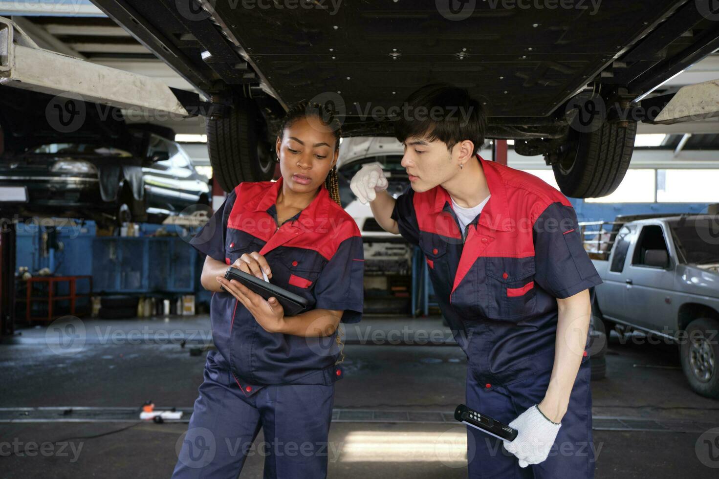 Two professional automotive worker partners discuss mechanical check undercarriage of lifted electric car, fix and repair at maintenance garage, and expert service business works industry occupation. photo