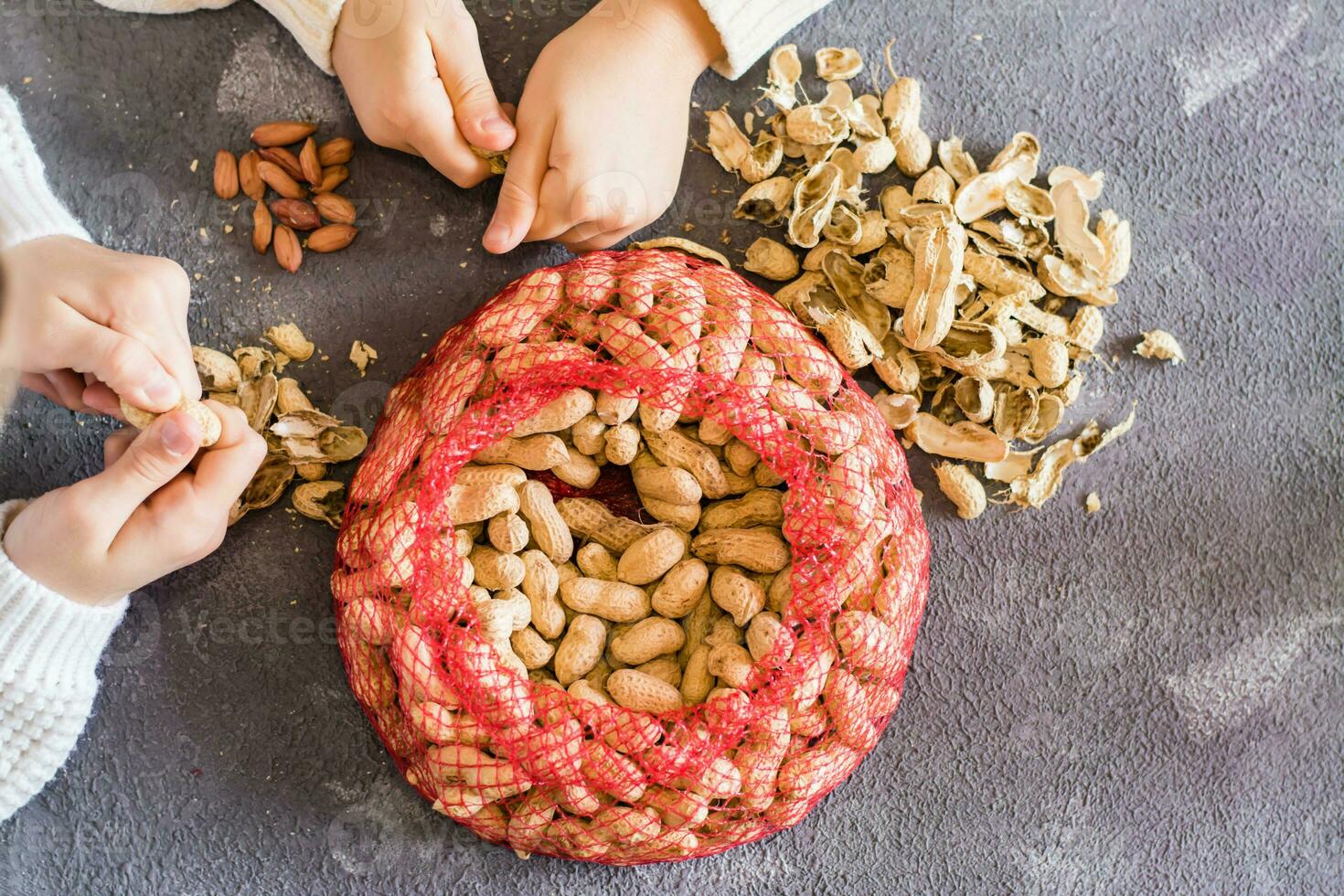 Peanut husks and a mesh bag with brown nuts on the table. Baby hands peeling nuts in the background. Lifestyle. Top view photo