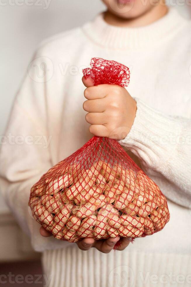 A girl in a cream sweater holds a mesh bag with peanuts in a shell. Lifestyle photo