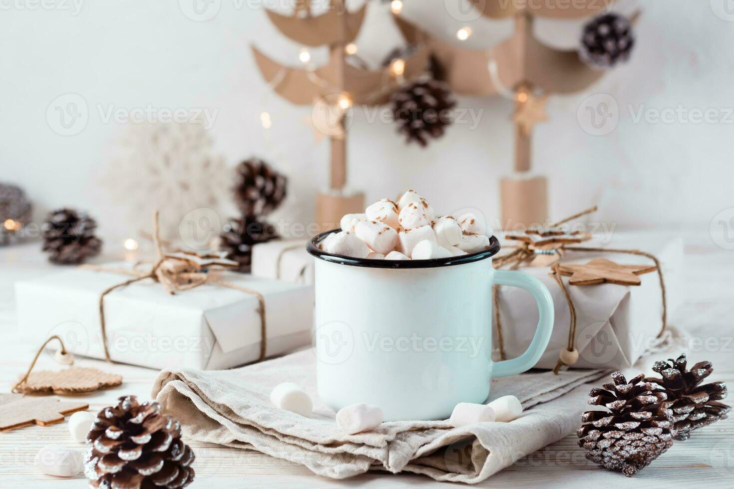 Cocoa and marshmallows in a mug next to craft gifts against the background of cardboard Christmas trees on a white wooden table. Hot sweet drinks photo