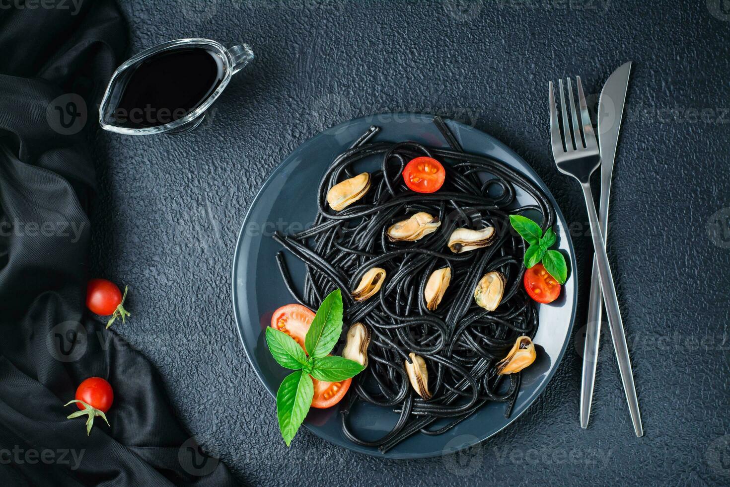 Ready-to-eat black spaghetti with mussels, tomatoes and basil on a plate on a black background. Food photography in dark colors. Top view photo