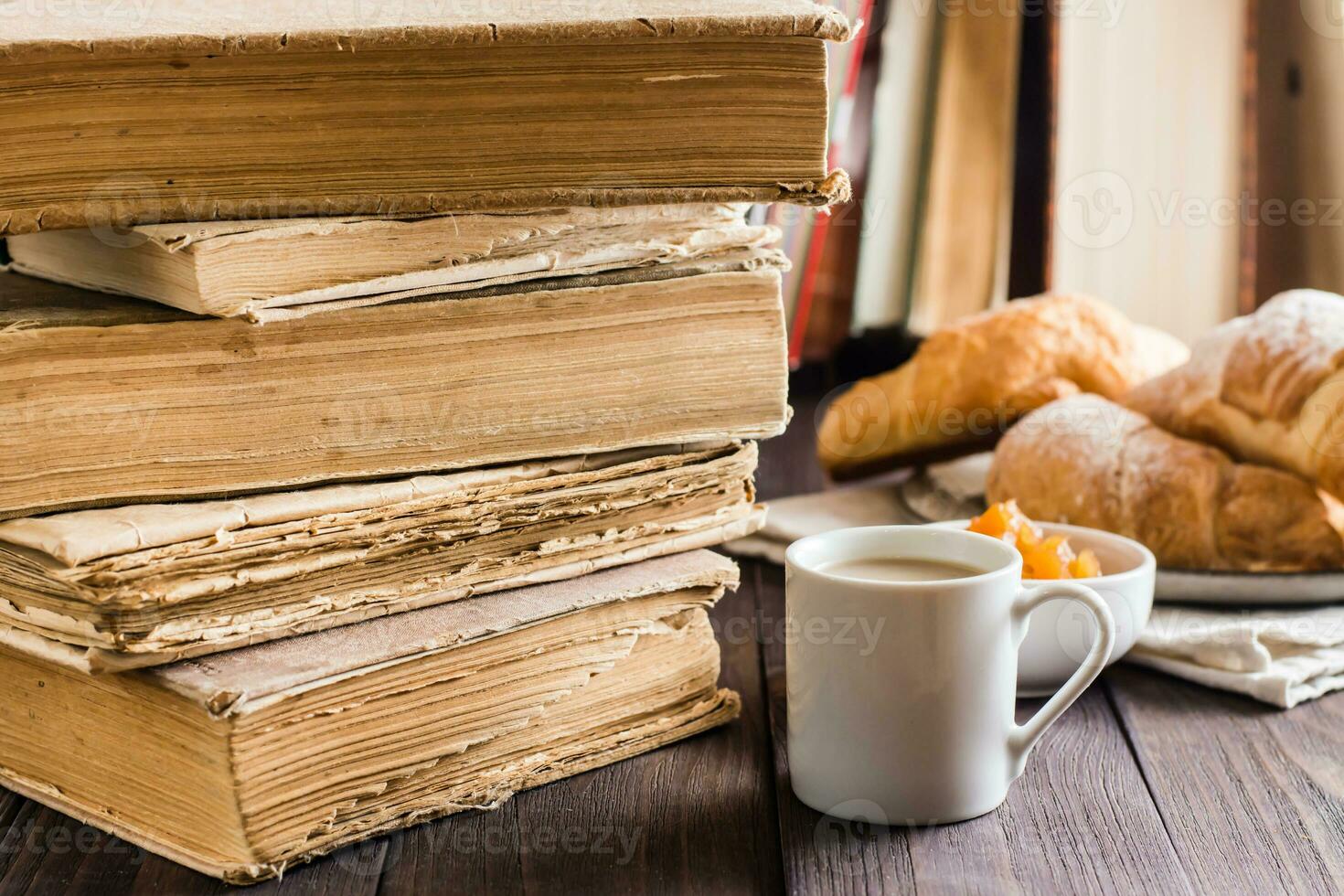 A cup of coffee and a plate with croissants in a vintage library with books on a wooden table. Relaxation, wellness and home reading. photo