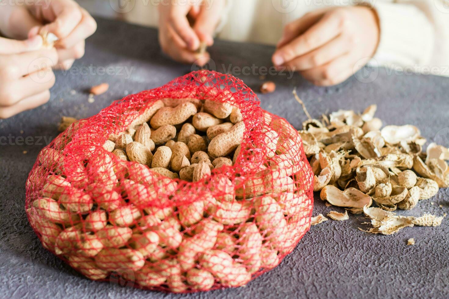Mesh bag with inshell peanuts and two girls in the background are peeling nuts at the table. Lifestyle photo