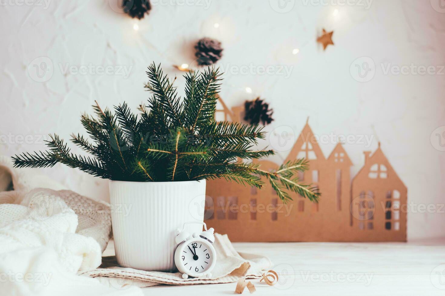Organization of a festive Christmas table. Fir bouquet in a vase and a white alarm clock on the background of a cardboard house on a white wooden table. Eco-friendly and homemade lifestyle photo