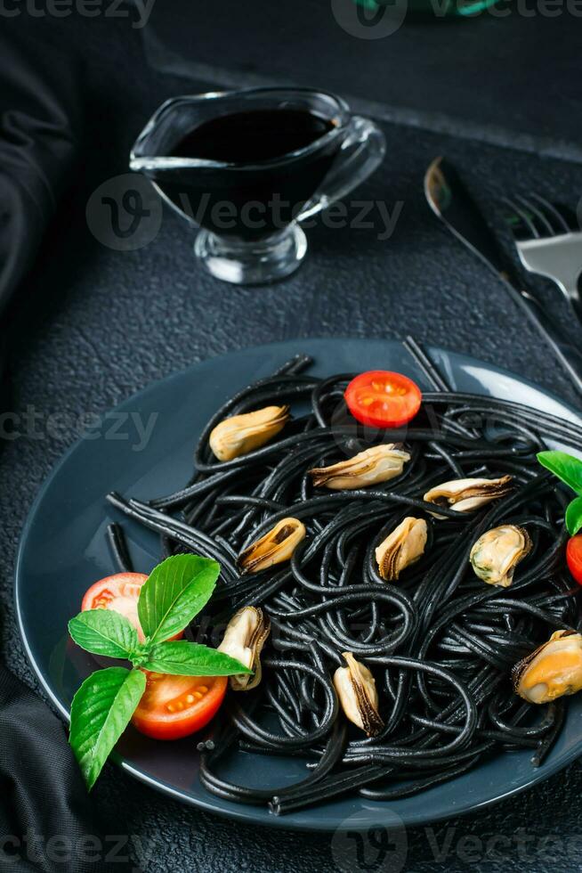 Ready to eat black spaghetti with mussels, tomatoes and basil on a plate and soy sauce on a black background. Food photography in dark colors. Close-up. Vertical view photo