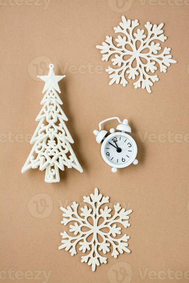 Concept Countdown on Christmas night. White alarm clock, decorative Christmas tree and snowflakes on beige cardboard. Top and vertical view photo