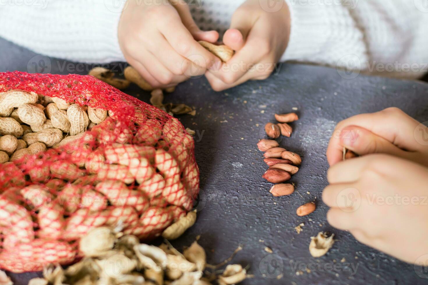 Peanut kernels, a mesh bag with unpeeled nuts and husks on the table. Children's hands are peeling nuts. Lifestyle photo