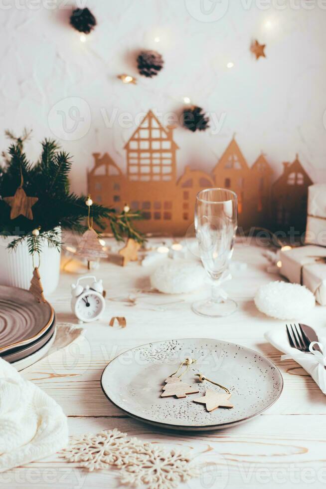 Organization of a festive Christmas table. Plate with wooden decorations, cutlery, champagne glass and cardboard house. Tablescapes on white wooden table. Vertical view photo