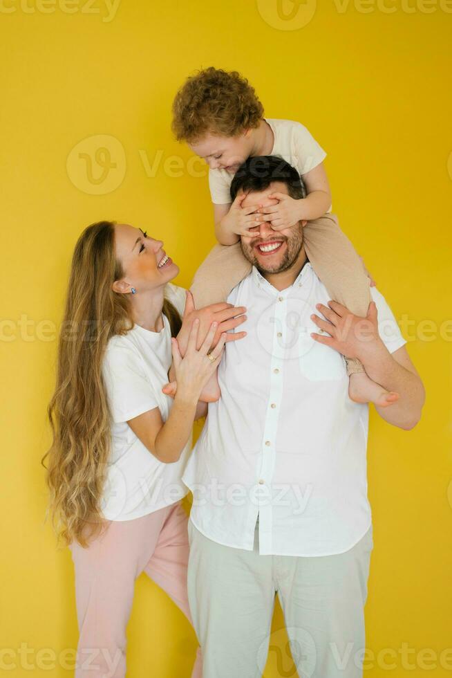 Happy parents hugging their son, expressing love, warm feelings. The child sits on his father's neck and covers his eyes with his palms. Having fun family isolated yellow background. photo