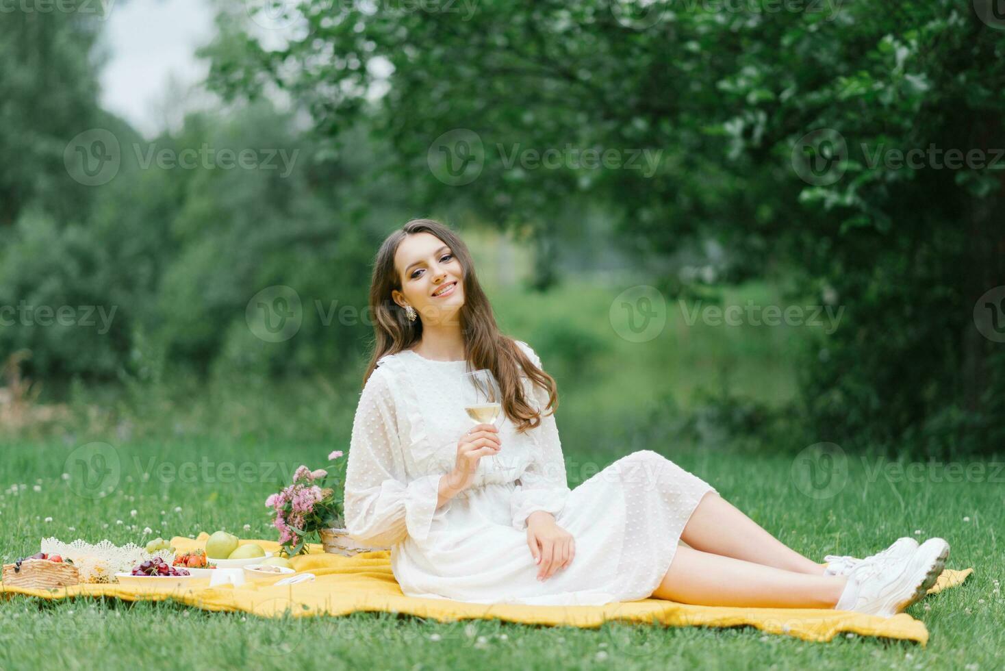 A happy smiling girl holds a glass of juice or wine and sits on a yellow blanket on a summer picnic outside the city photo