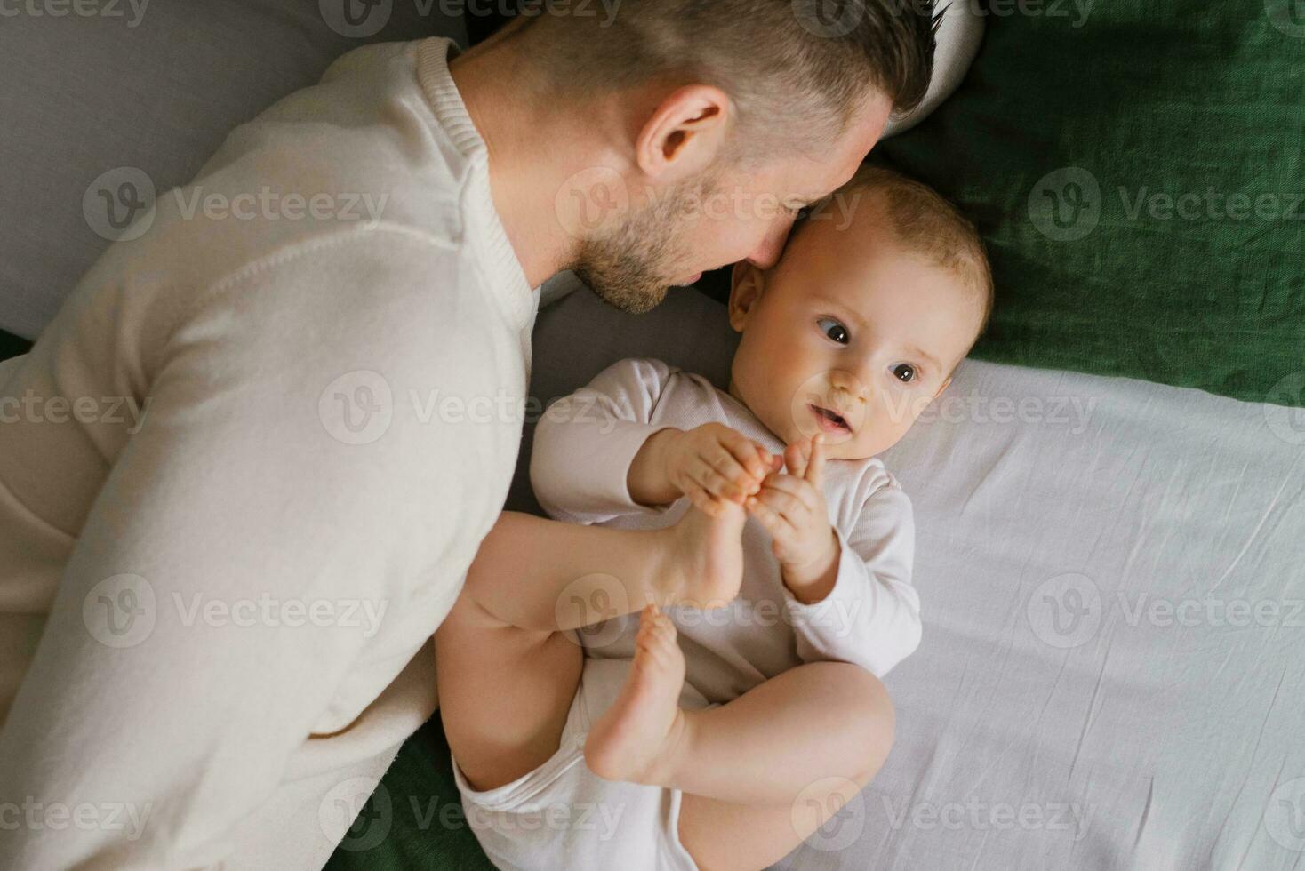 Cheerful, beautiful newly-made dad and a charming six-month-old baby lie together on green and white underwear. A young father with a small child in a home portrait. Top view photo