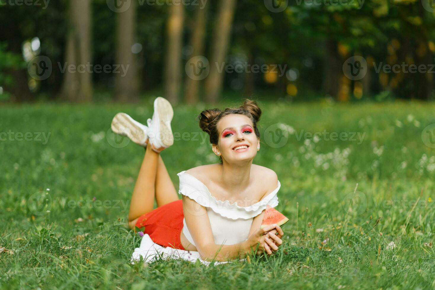 un contento joven mujer con brillante maquillaje sostiene un pedazo de sandía en su manos y sonrisas al aire libre en verano, acostado en un cobija foto